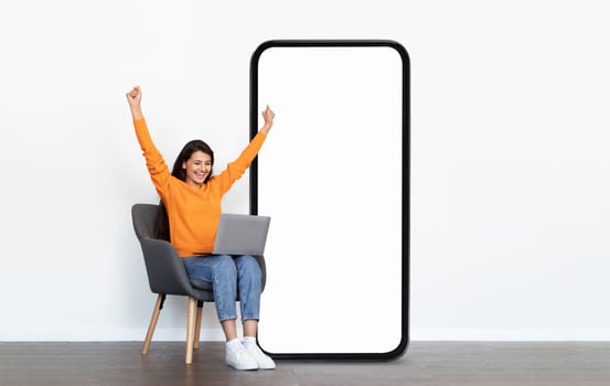 A young woman in an orange sweater sits on a chair, excitedly raising her arms in victory while using a laptop next to an oversized blank phone screen, symbolizing achievement and technology. photo
