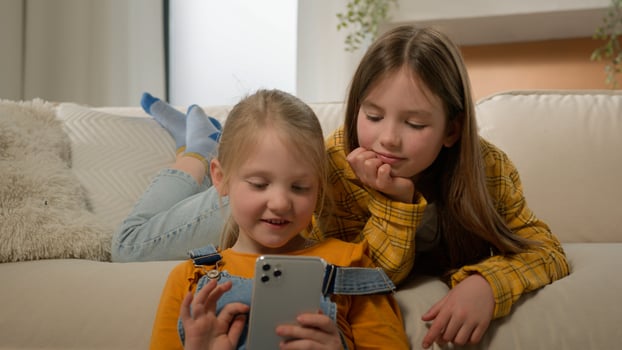 Two funny happy caucasian kids girls sisters friends children having fun playing on phone in living room couch play mobile online game together using smartphone browsing social network in Internet photo
