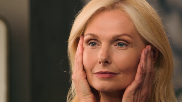 Close up face Caucasian beautiful woman middle aged mature 50s old lady happy female beauty looking at mirror reflection smile touch face perfect moisturizing skin anti-aging lifting therapy collagen photo