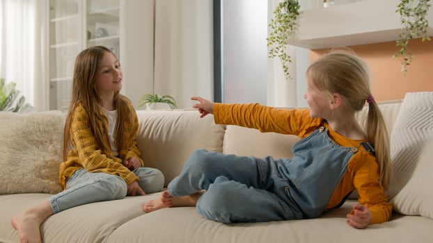 Two laughing little Caucasian children friends kids girls playing couch pointing finger gesture you sisters relationship daughters family fun talking contend debate sharing sofa together living room photo