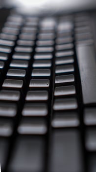 A close-up of a sleek black keyboard with square keys, resting on a dark, textured surface. The keyboard's sharp angles and minimalist design create a modern photo