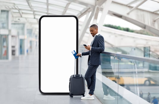 African American businessman stands casually in an airport terminal, holding a smartphone while resting a suitcase nearby. The terminal features modern architecture and a blank digital display. photo