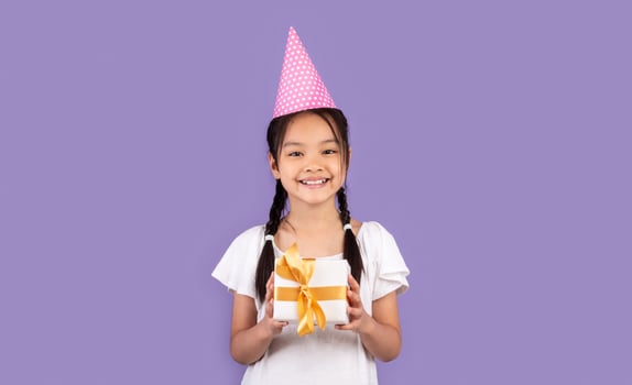 Happy Birthday. Asian Girl Holding B-Day Gift Box Wearing Pink Party Hat Posing Over Purple Background In Studio. Free Space photo