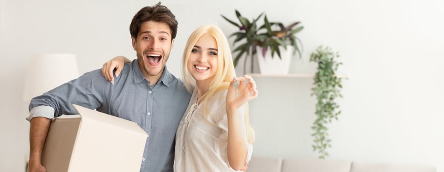 A joyful couple poses in their new living room, holding a cardboard box and smiling broadly. They are surrounded by plants and a bright, welcoming atmosphere, indicating a fresh start, copy space photo