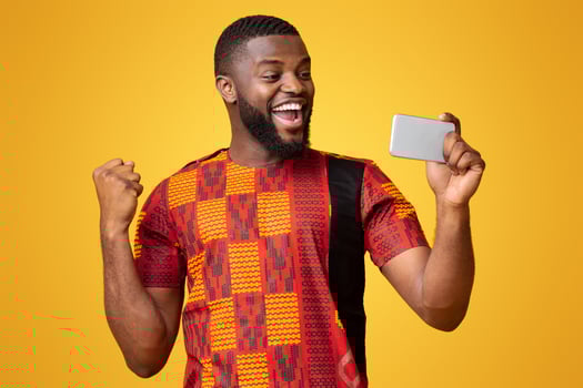 Happy black man in african t-shirt holding phone and expressing happiness, shouting and clenching fist, yellow background photo