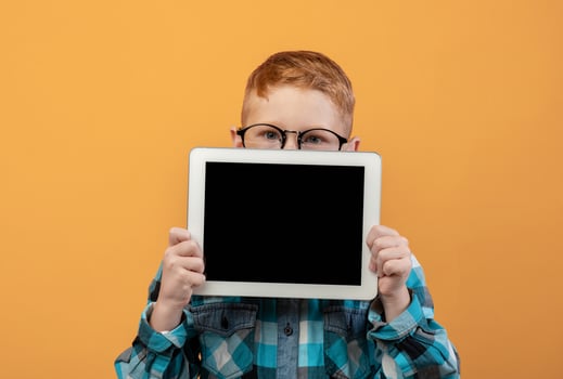 Ginger boy in eyeglasses hiding behind digital tablet with empty screen over yellow studio background. Cute redhead schoolboy showing pad, mockup, advertisement for cute application photo