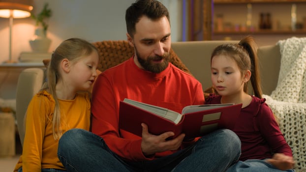 Happy Caucasian family father children little school girls daughters read book together single parent babysitter dad adopted kids siblings smile talk watch pictures photo album at evening home weekend