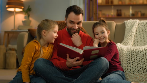 Caucasian family late night evening at home parent dad father man reading book kids watching photo album funny pictures happy cheerful amazed children little girls daughters schoolgirls enjoy learning
