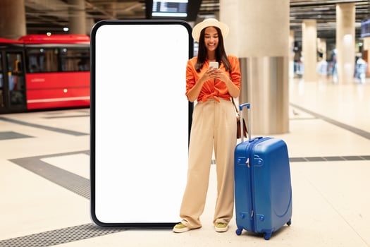 In a bustling airport, a young woman stands beside a large blank advertisement display while checking her phone. She holds a suitcase, appearing ready to embark on her journey. photo