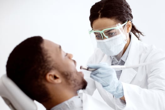 Black Dentist Lady In Protective Medical Mask Treating Male Patient's Teeth With Dental Drill, Professional Stomatologist Using Sterile Tools During Medical Procedure In Modern Clinic, Closeup Shot photo
