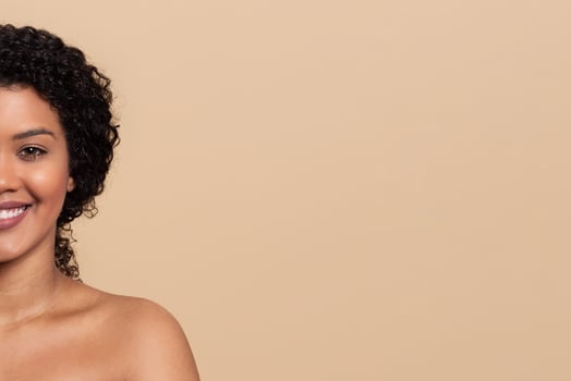 A woman with curly hair smiles confidently, showcasing her radiant complexion. The soft lighting enhances her features, creating a warm and inviting atmosphere in the background, blank space photo