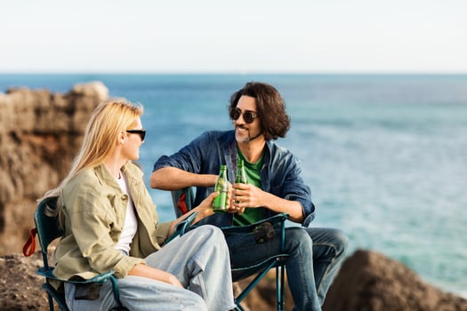 A man and woman relax on folding chairs near the ocean, sipping beverages and having a cheerful conversation on a sunny afternoon. The beautiful coastline enhances their moment together. photo