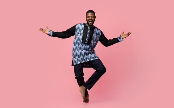 Full size photo of friendly black man dancing over pink studio background, copy space. Cheerful african american guy wearing traditional costume and showing welcoming gesture, studio shot