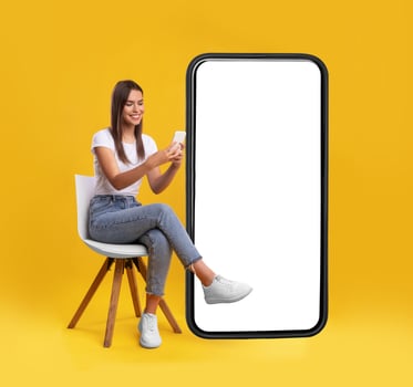 Cheerful smiling female sitting on chair near big cell phone with empty white screen, using mobile device, checking new cool app on orange studio background, mock up for website or application design photo