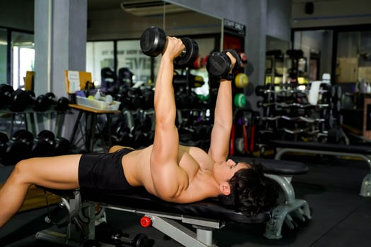 Closeup Asian handsome man with perfect body playing dumbbells' weight training in the lying position to build shoulder and chest muscles at fitness center. photo