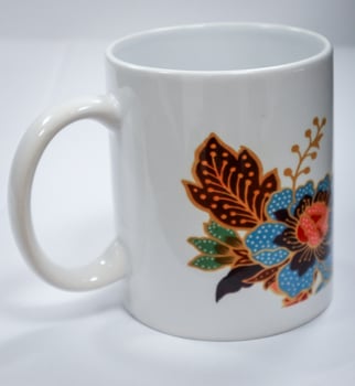 A white mug with a flower and leaf design using Indonesian batik motifs. The background is plain white with no people photo