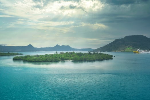 Serene tropical landscape with a lush green island, turquoise waters, and distant mountains under a cloudy sky with sunrays. A yellow ferry crosses the calm bay, inviting travel and adventure. photo