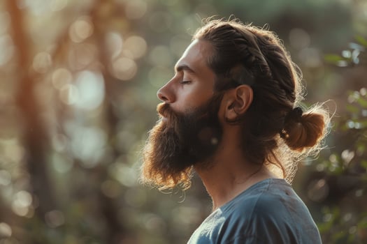 A man with a beard and long hair is sitting in the woods, looking to his right photo