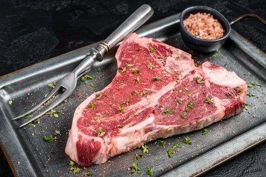 T-bone steak, raw marbled beef meat in a steel kitchen tray with herbs. Black background. Top view photo