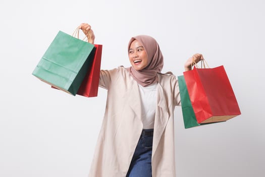 Portrait of excited Asian hijab woman in casual suit holding and showing colorful paper shopping bags. Shopaholic girl and discount buying concept. Isolated image on white background photo