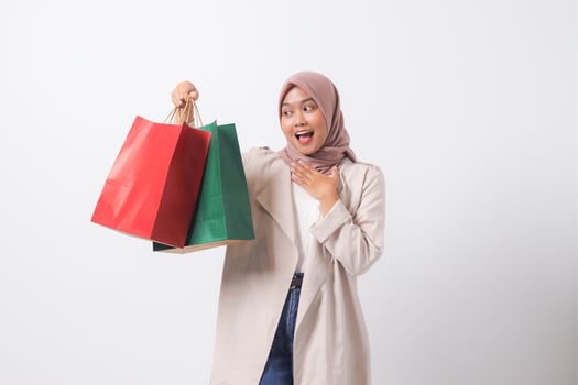 Portrait of shocked Asian hijab woman in casual suit holding paper shopping bags while covering mouth. Shopaholic girl and discount buying concept. Isolated image on white background photo