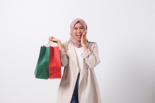 Portrait of shocked Asian hijab woman in casual suit holding paper shopping bags while covering mouth. Shopaholic girl and discount buying concept. Isolated image on white background photo