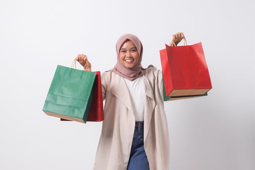 Portrait of excited Asian hijab woman in casual suit holding and showing colorful paper shopping bags. Shopaholic girl and discount buying concept. Isolated image on white background photo