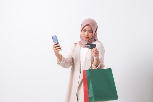 Portrait of excited Asian hijab woman in casual suit holding shopping bags and credit card while using mobile phone. Shopaholic girl and discount buying concept. Isolated image on white background photo