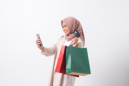 Portrait of excited Asian hijab woman in casual suit holding credit card and shopping bags while using mobile phone. Shopaholic girl and discount buying concept. Isolated image on white background photo