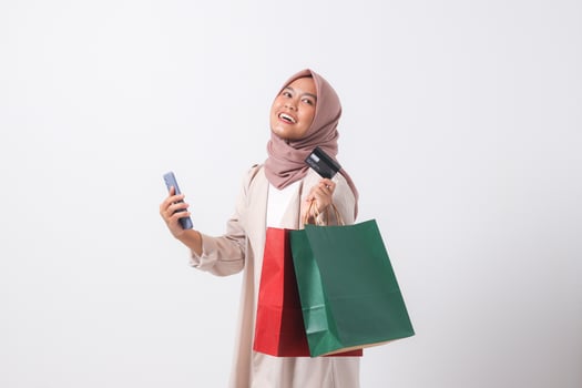 Portrait of excited Asian hijab woman in casual suit holding credit card and shopping bags while using mobile phone. Shopaholic girl and discount buying concept. Isolated image on white background photo