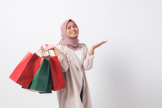 Portrait of excited Asian hijab woman in casual suit holding paper shopping bags while pointing with finger. Shopaholic girl and discount buying concept. Isolated image on white background photo