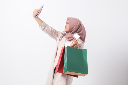 Portrait of excited Asian hijab woman in casual suit holding credit card and shopping bags while taking selfie with mobile phone. Shopaholic girl concept. Isolated image on white background photo