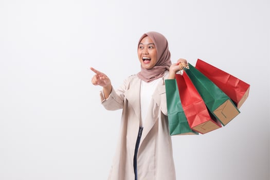 Portrait of excited Asian hijab woman in casual suit holding paper shopping bags while pointing with finger. Shopaholic girl and discount buying concept. Isolated image on white background photo