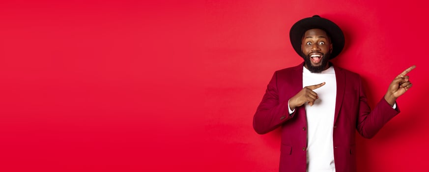 Winter holidays and shopping concept. Stylish african american man pointing fingers right at copy space for logo, standing over red background photo