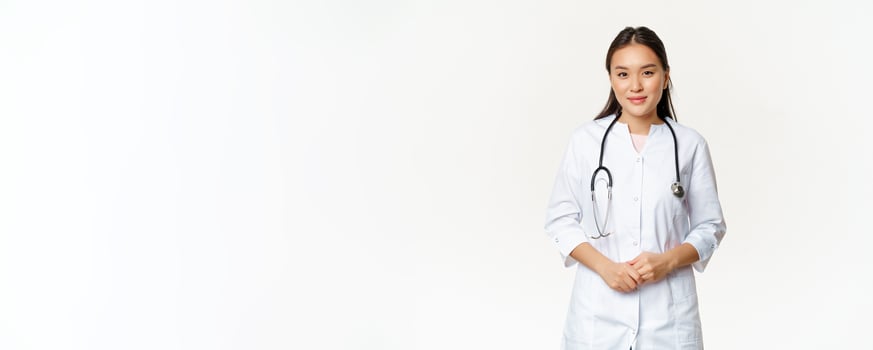 Smiling asian medical worker with stethoscope, wearing doctor uniform, looking helpful at patient, standing over white background photo