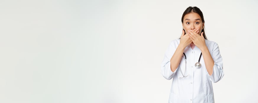 Image of shocked female asian doctor covers her lips with hands, close mouth and looking worried, taboo gesture, standing over white background photo