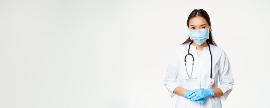 Covid-19 and healthcare concept. Young asian female doctor in medical mask, rubber gloves and clinic uniform looking ready to help, listening patient, white background photo