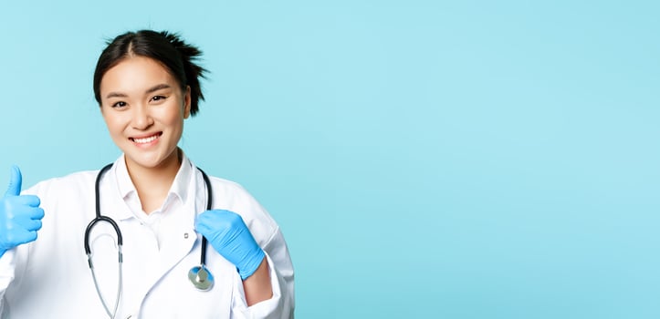 Smiling asian woman therapist, physician with stethoscope shows thumbs up, standing over blue background photo