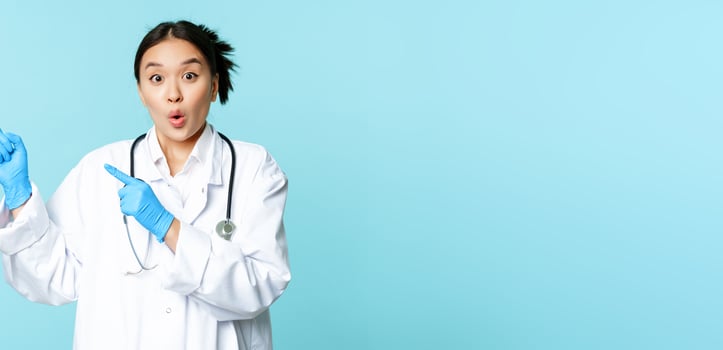 Surprised and enthusiastic asian doctor, female medical worker pointing fingers at upper left corner, showing disocunt, standing over blue background photo