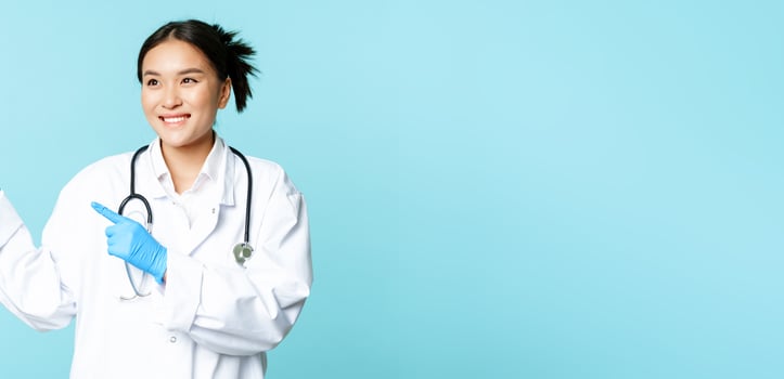 Asian smiling doctor, medical healthcare worker, pointing fingers left, looking pleased at advertisement, advertising product, blue background photo