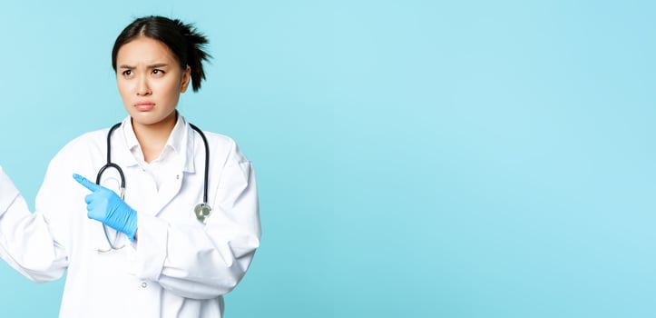 Angry asian medical worker, nurse or doctor express disapproval, pointing left and furrow eyebrows, standing over blue background photo