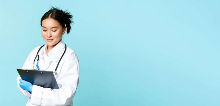 Asian female nurse or doctor writing down on clipboard with pen, smiling while examining patients, standing in uniform over blue background photo
