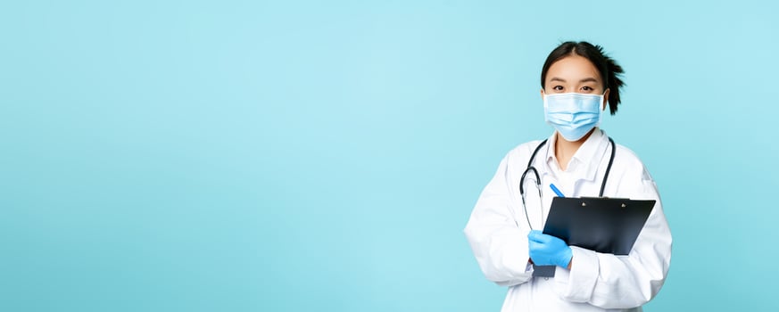 Covid-19 and quarantine concept. Smiling asian female physician, nurse in face mask holding clipboard, shift at hospital, standing over blue background photo