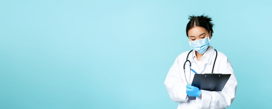 Image of korean doctor, female nurse in medical face mask, writing down patient information, examining on hospital shift, standing in uniform over blue background photo