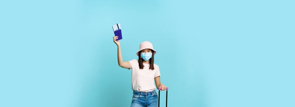 Full length of happy asian tourist, girl travelling abroad, posing with her suitcase in medical face mask, showing passport and tickets, blue background photo