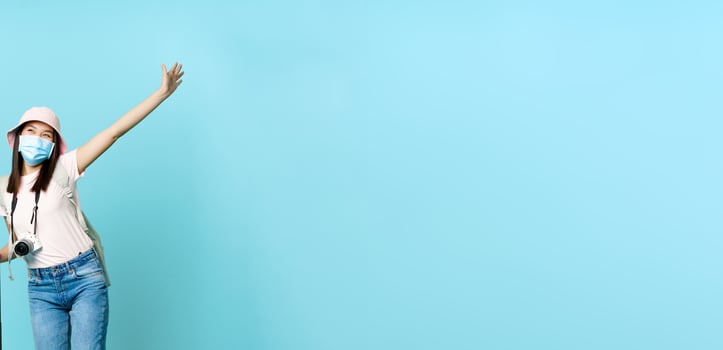 Happy asian girl in medical mask, tourist posing excited with suitcase, going on vacation, travelling abroad, standing over blue background photo