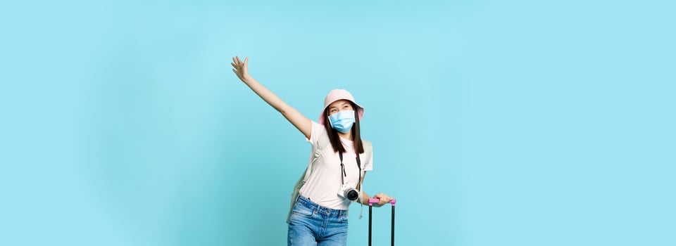Happy asian girl in medical mask, tourist posing excited with suitcase, going on vacation, travelling abroad, standing over blue background photo