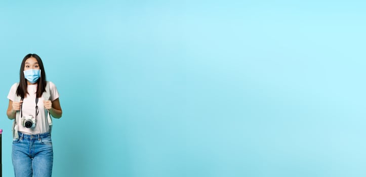 Enthusiastic girl traveller, asian tourist with backpack and suitcase, wearing medical face mask, excited about journey, vacation trip, standing over blue background photo