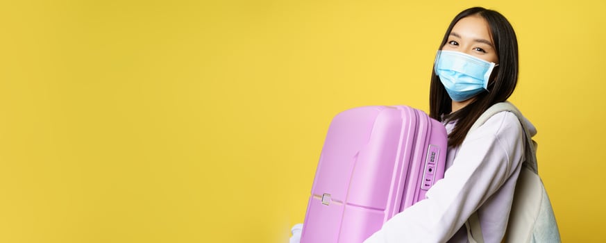 Close up of happy asian girl tourist in medical face mask, holding pink cute suitcase, going on vacation, travelling during pandemic, yellow background photo