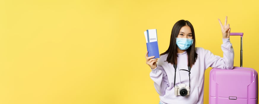 Japanese girl in medical mask, showing her passport and flight travel pass tickets, peace sign, posing with suitcase, going on vacation, yellow background photo